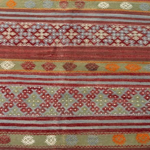 May include: A rectangular, hand-woven rug with a geometric pattern. The rug features alternating bands of red, gray, and green, with intricate diamond and floral motifs in shades of red, orange, and white. The rug's texture is visible.