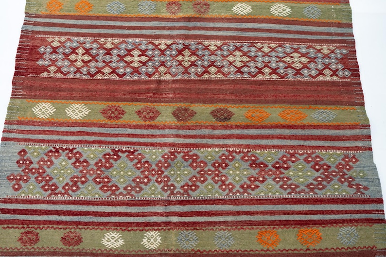 May include: A rectangular, hand-woven rug with a geometric pattern. The rug features alternating bands of red, gray, and green, with intricate diamond and floral motifs in shades of red, orange, and white. The rug's texture is visible.