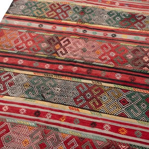 May include: A vibrant, patterned rug with a mix of geometric and linear designs. The rug features a rich color palette of red, green, black, and cream, with intricate patterns and stripes. The rug's design is a blend of traditional and contemporary styles.