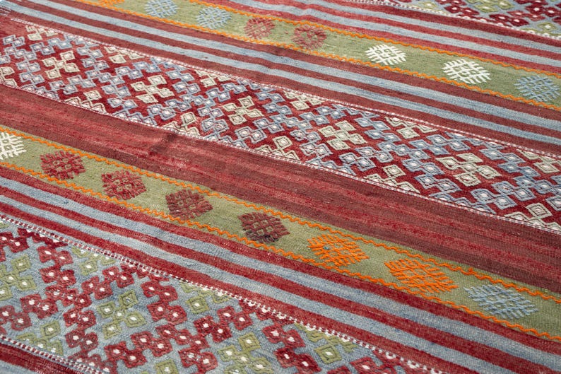 May include: A colorful rug with a geometric pattern. The rug features alternating stripes of red, blue, and green, with diamond and floral motifs. The rug's design incorporates various shades of red, blue, green, and orange.