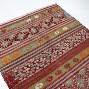 May include: A rectangular rug with a geometric pattern. The rug features alternating stripes of red, green, and gray, with diamond and floral motifs. The rug has a fringed edge and is suitable for home decor.