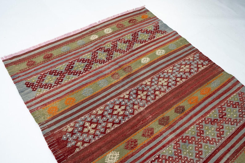 May include: A rectangular rug with a geometric pattern. The rug features alternating stripes of red, green, and gray, with diamond and floral motifs. The rug has a fringed edge and is suitable for home decor.