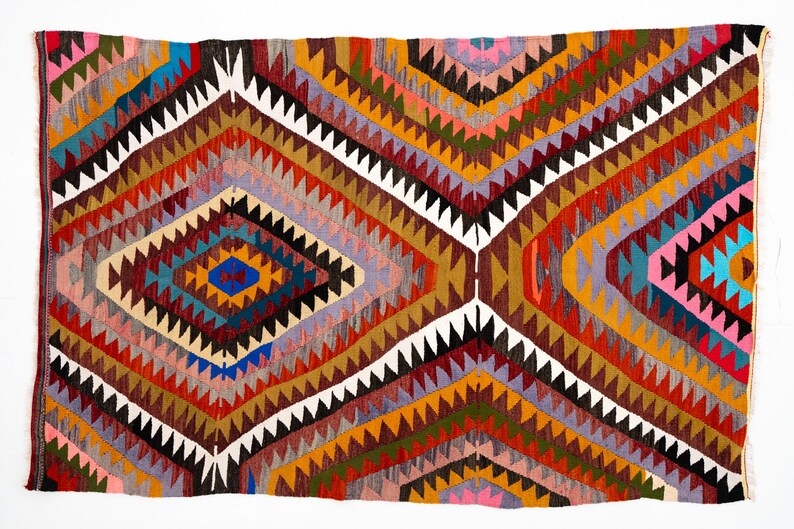 K&ouml;nnte beinhalten: Ein rechteckiger, handgewebter Teppich mit geometrischem Design. Der Teppich weist ein Rautenmuster in Rot-, Orange-, Gelb-, Braun-, Blau- und Wei&szlig;t&ouml;nen auf. Die Kanten sind mit einem wei&szlig;en Rand versehen. Der Teppich ist wahrscheinlich aus Wolle oder einem &auml;hnlichen Material.