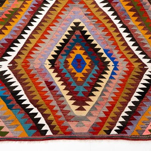 K&ouml;nnte beinhalten: Ein farbenfroher, geometrischer Teppich mit einem Rautenmuster. Der Teppich zeigt eine zentrale Raute in Blau-, Orange- und Gelbt&ouml;nen, umgeben von sich wiederholenden Zickzackmustern in verschiedenen Farben. Der Teppich besteht aus gewebtem Stoff.