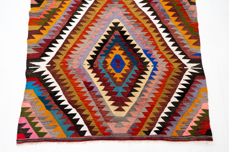 K&ouml;nnte beinhalten: Ein farbenfroher, geometrischer Teppich mit einem Rautenmuster. Der Teppich zeigt eine zentrale Raute in Blau-, Orange- und Gelbt&ouml;nen, umgeben von sich wiederholenden Zickzackmustern in verschiedenen Farben. Der Teppich besteht aus gewebtem Stoff.