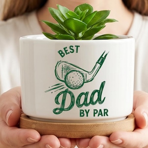 Pode incluir: Um vaso de cerâmica branco com uma planta suculenta verde. O vaso tem o texto "BEST Dad BY PAR" com um gráfico de taco de golfe e bola. O vaso assenta sobre um pires redondo de madeira.