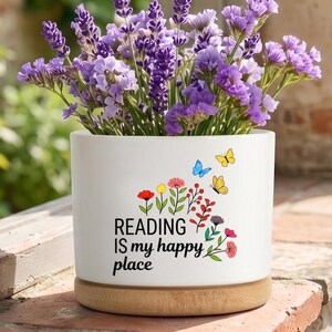 Pode incluir: Um vaso cilíndrico de cerâmica branca com base de madeira clara, preenchido com flores de lavanda roxa e statice. A frente apresenta um gráfico floral colorido com borboletas e o texto preto "READING IS my happy place" impresso.