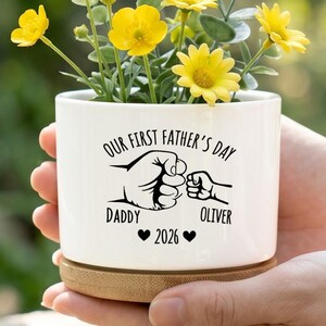 May include: A white ceramic planter with yellow flowers, featuring the text "OUR FIRST FATHER'S DAY" and the names "DADDY" and "OLIVER" with a fist bump graphic. The planter sits on a wooden base.
