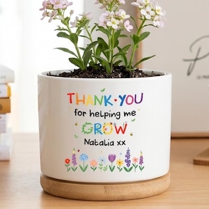 Puede incluir: Una maceta cilíndrica de cerámica blanca con base de bambú que contiene una pequeña planta con flores de color rosa claro y blanco. La maceta presenta el texto colorido "THANK YOU for helping me GROW" y "Natalia xx" sobre ilustraciones florales.