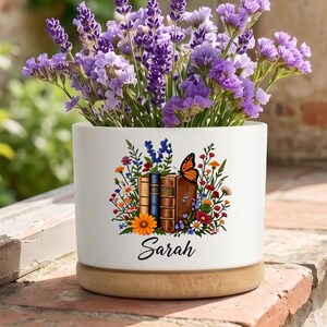 Pode incluir: Um vaso cilíndrico de cerâmica branca com base de madeira, preenchido com flores de lavanda roxas. A frente apresenta um gráfico colorido de livros empilhados, flores silvestres e uma borboleta monarca, com o nome Sarah em preto.