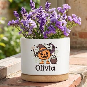May include: A white cylindrical ceramic planter with a wooden base, filled with purple lavender flowers. The front features a graphic of a cartoon pumpkin wearing a witch hat, holding a wand next to a cauldron, with the name Olivia printed below.