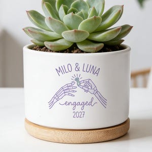 May include: A white ceramic cylindrical planter with a light wood base containing a green succulent. The planter features a purple graphic of two skeleton hands exchanging a ring with the text "MILO & LUNA engaged 2027" printed in purple.