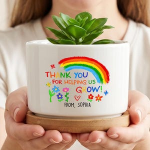 Può includere: Un vaso cilindrico in ceramica bianca con base in legno, contenente una pianta succulenta verde. Il vaso presenta una grafica colorata con arcobaleno, piccole illustrazioni di fiori e il testo: THANK YOU FOR HELPING US GROW! FROM, SOPHIA.