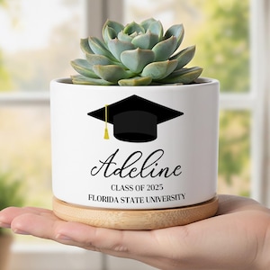 Op de afbeelding: Een witte keramische plantenbak met een vetplant, een afstudeerhoed-afbeelding en de naam "Adeline" met de tekst "Class of 2025" en "Florida State University". De plantenbak staat op een houten voet.