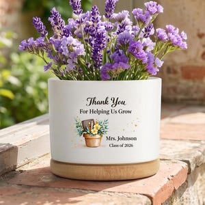 Puede incluir: Una maceta cilíndrica de cerámica blanca con base de madera clara, llena de flores de lavanda moradas. El frente presenta el texto negro Thank You For Helping Us Grow, una pequeña ilustración de una planta, Mrs. Johnson y Class of 2026.