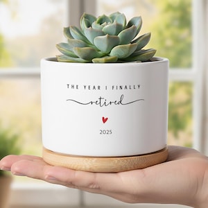 Könnte beinhalten: Ein weißer Keramik-Pflanztopf mit einer Sukkulentenpflanze, die auf einem Holzuntersetzer ruht und in einer Hand gehalten wird. Der Topf trägt den Text "THE YEAR I FINALLY retired" mit einem roten Herz und dem Jahr 2025.