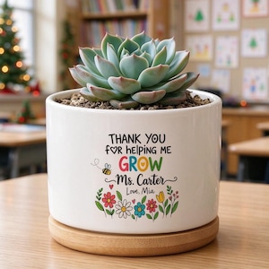May include: A white ceramic cylindrical planter with a bamboo base containing a green succulent. The planter features the text "THANK YOU FOR helping me GROW Ms. Carter Love, Mia" with a small bee and colourful flower illustrations.