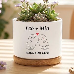 May include: A white cylindrical ceramic planter sitting on a round wooden base. The planter features a graphic of two ghosts holding hands with a red heart above them, the text Leo + Mia, and the phrase BOOS FOR LIFE. Green succulents are planted inside.