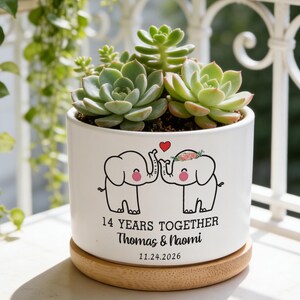 Puede incluir: Maceta de cerámica blanca con base de madera, con un arreglo de suculentas. La maceta está decorada con dos elefantes de dibujos animados tocándose las trompas, un corazón y el texto "14 YEARS TOGETHER" con nombres y una fecha.