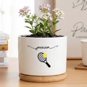 May include: A white cylindrical ceramic planter with a bamboo saucer base. The planter features the name Jessica in black cursive script above a black tennis racket graphic with a yellow tennis ball. A small plant with light pink flowers sits inside.