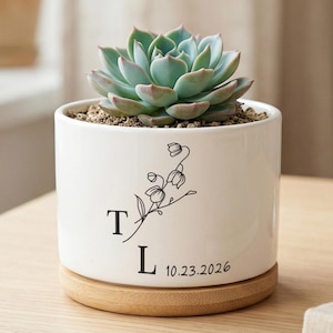 May include: A white ceramic planter with a succulent plant. The planter has a wooden base and features a black floral design with the letters "T" and "L" and the date "10.23.2026".