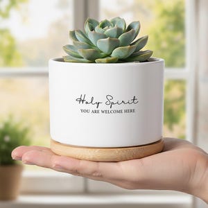 May include: A white ceramic plant pot with the text "Holy Spirit, You Are Welcome Here" in black script. The pot contains a green succulent and rests on a small wooden saucer. The pot is held in a person's hand.