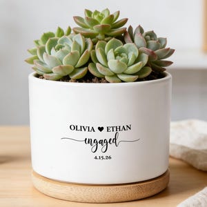以下が含まれることがあります： 多肉植物が入った白いセラミックプランター。 プランターは木製のベースで、「Olivia Ethan engaged 4.15.26」というテキストが黒で表示されています。 背景には木製の鹿の置物があります。