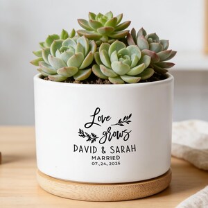 May include: White ceramic planter with a succulent plant. The planter has the text "Love grows" with the names "David & Sarah" and the wedding date "07.24.2026". The planter sits on a wooden base.