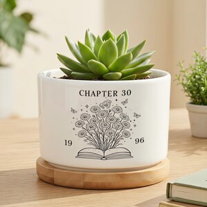 May include: A white ceramic cylindrical planter resting on a circular bamboo saucer. The planter features a black line art graphic of an open book with a bouquet of daisies and butterflies, with the text "CHAPTER 30" above and "19 96" below.