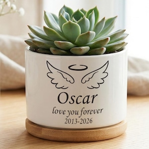 Może przedstawiać: Biała ceramiczna doniczka z sukulentem, skrzydłami anioła i imieniem "Oscar" z tekstem "love you forever 2013-2026". Doniczka stoi na drewnianej podstawce.