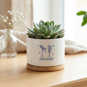 Peut inclure: Un pot de fleurs en céramique blanche avec une plante succulente verte, posé sur un dessous de plat en bois. Le pot présente un motif floral bleu et le texte "It takes a long time to grow an old friend."
