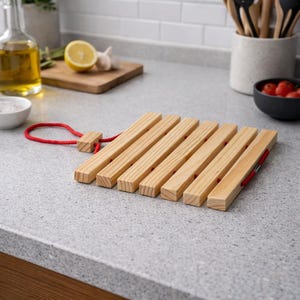 May include: Wooden trivet with six parallel wooden slats, connected by red rope. The trivet is made of light-coloured wood and has a red loop for hanging. Kitchenware.