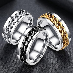 Peut inclure: Trois bagues en acier inoxydable avec un motif de chaîne. Une bague a une chaîne argentée, une chaîne noire et la troisième une chaîne dorée. Les bagues sont présentées sur une surface gris foncé.