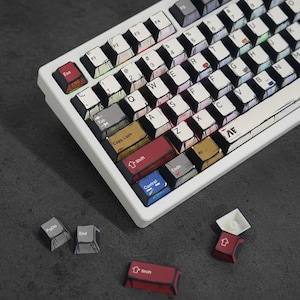 Puede incluir: Un teclado mecánico blanco con teclas personalizadas. Las teclas presentan un estilo dibujado a mano con diversos colores y diseños. Varias teclas están separadas, mostrando los componentes del teclado.