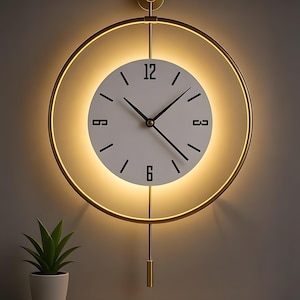 May include: A modern wall clock with a white face, black numerals, and black hands. The clock is surrounded by a gold-colored circular frame with an integrated warm light. A gold pendulum hangs below. A small potted plant sits on a wooden shelf below the clock.