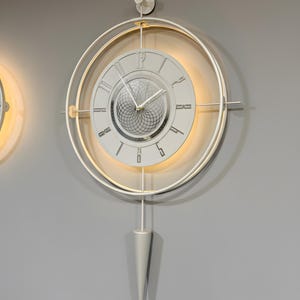 Pode incluir: Um relógio de parede branco com um design circular. O relógio apresenta algarismos romanos e um padrão interno decorativo. Possui um pêndulo e é rodeado por uma luz brilhante.