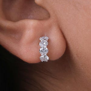 Könnte beinhalten: Nahaufnahme eines kleinen Creolen-Ohrrings aus Weißgold mit fünf klaren, rund geschliffenen Edelsteinen. Der Ohrring wird an einem Ohr getragen, wobei der Fokus auf dem Glanz und dem Design des Schmucks liegt.
