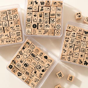 Puede incluir: Cuatro recipientes de plástico transparente llenos de sellos de madera con varias ilustraciones en blanco y negro. Los sellos incluyen iconos, emojis y frases cortas. Varios sellos sueltos están dispersos.