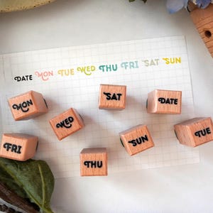 May include: A set of eight light-coloured wooden square blocks, each featuring a black stamped abbreviation for days of the week including MON, TUE, WED, THU, FRI, SAT, SUN, and the word DATE, arranged on a white grid paper with colourful text.