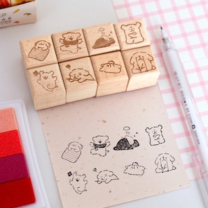 Op de afbeelding: Een set van acht houten stempels met schattige hondenillustraties. De stempels zijn gerangschikt op een lichtgekleurd oppervlak, naast een vel papier met gestempelde hondenontwerpen. Een witte pen en een veelkleurig inktkussen zijn ook aanwezig.