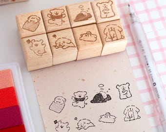 May include: A set of eight wooden stamps with cute dog illustrations. The stamps are arranged on a light-colored surface next to a sheet of paper with stamped dog designs. A white pen and a multi-colored ink pad are also present.