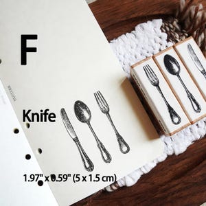 May include: Three rectangular wooden stamps featuring detailed black ink illustrations of a knife, a spoon, and a fork. The stamps are labeled with the letter F and the word Knife, with dimensions of 1.97 inches by 0.59 inches printed on the paper.