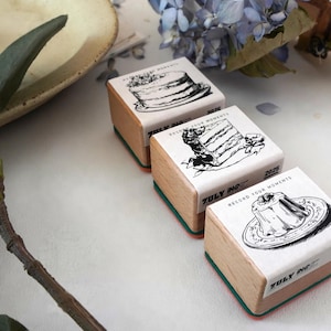 May include: Three wooden rubber stamps with light wood blocks and colorful bases. Each stamp features a black and white line drawing of a dessert, including a whole cake, a slice of cake, and a pudding, with the text ZULY INC and RECORD YOUR MOMENTS 2025.