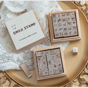 以下が含まれることがあります： 蓋にEMOJI STAMPと記された木製とゴム製のスタンプ25個セット。コレクションには、天気記号、旅行アイコン、日常のオブジェクトなど、黒インクで描かれた様々なアイコンが刻印された正方形のスタンプが入った2つの木製トレイが含まれています。