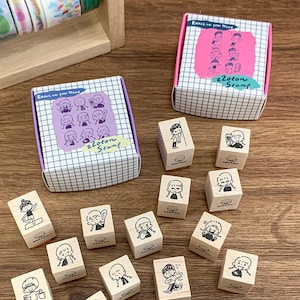 Könnte beinhalten: Holzstempel mit verschiedenen Cartoon-Illustrationen, darunter ein Mädchen mit Zöpfen, werden auf einer Holzoberfläche präsentiert. Zwei Boxen mit dem Text "Reach into your Heart" und "zzoton Stamp" sind ebenfalls zu sehen. Die Boxen haben ein Gittermuster.