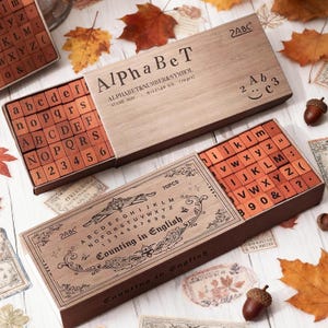 Könnte beinhalten: Holzstempel-Sets mit Alphabet und Zahlen. Die Sets enthalten Groß- und Kleinbuchstaben, Zahlen und Symbole. Die Boxen sind aus hellbraunem Holz gefertigt und tragen die Aufschrift "AlphaBeT" und "Counting in English".