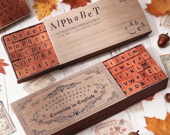 Puede incluir: Juegos de sellos de alfabeto y números de madera. Los juegos incluyen letras mayúsculas y minúsculas, números y símbolos. Las cajas son de madera marrón claro y presentan el texto "AlphaBeT" y "Counting in English".