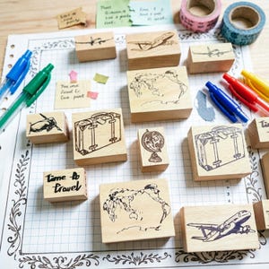 May include: A collection of wooden rubber stamps featuring travel-themed illustrations including airplanes, suitcases, a globe, a map, and a hand writing. The stamps are arranged on a grid-patterned paper alongside colorful pens and washi tape.