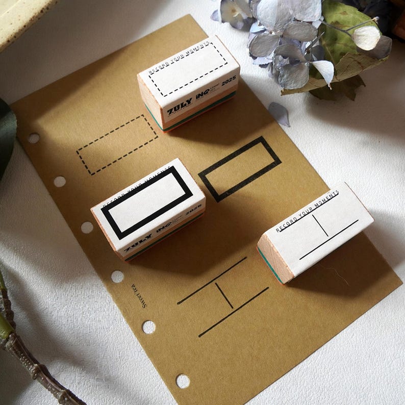 May include: Three rectangular wooden rubber stamps with white tops and green bases. The stamps feature black ink designs including a dashed rectangle, a solid rectangle, and a horizontal line with a vertical divider, all labeled with the text RECORD YOUR MOMENTS and ZULY INC 2025.