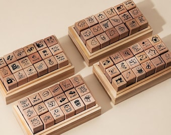 May include: Four sets of wooden stamps with various hand-drawn icons and words, including a camera, coffee cup, and the word "test." Each set is arranged in a wooden tray, ideal for crafting or journaling.